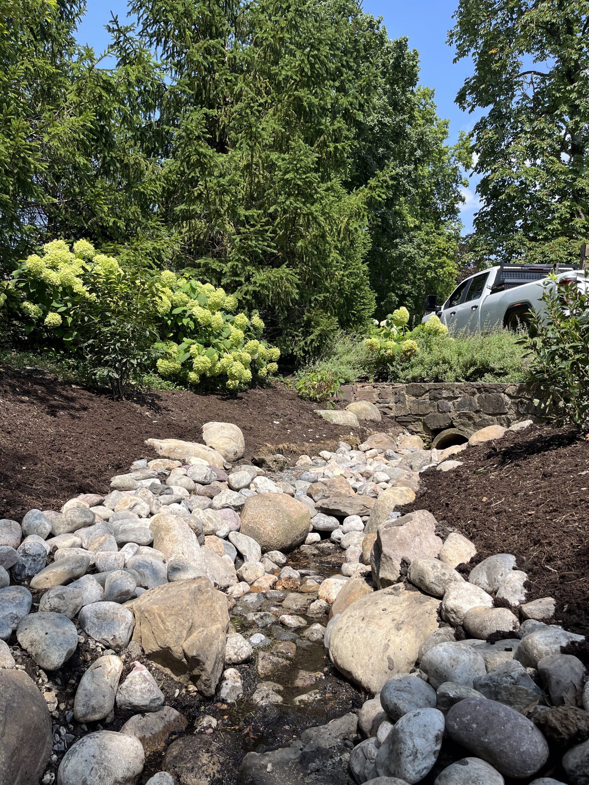 Rocky garden stream with lush greenery and car.