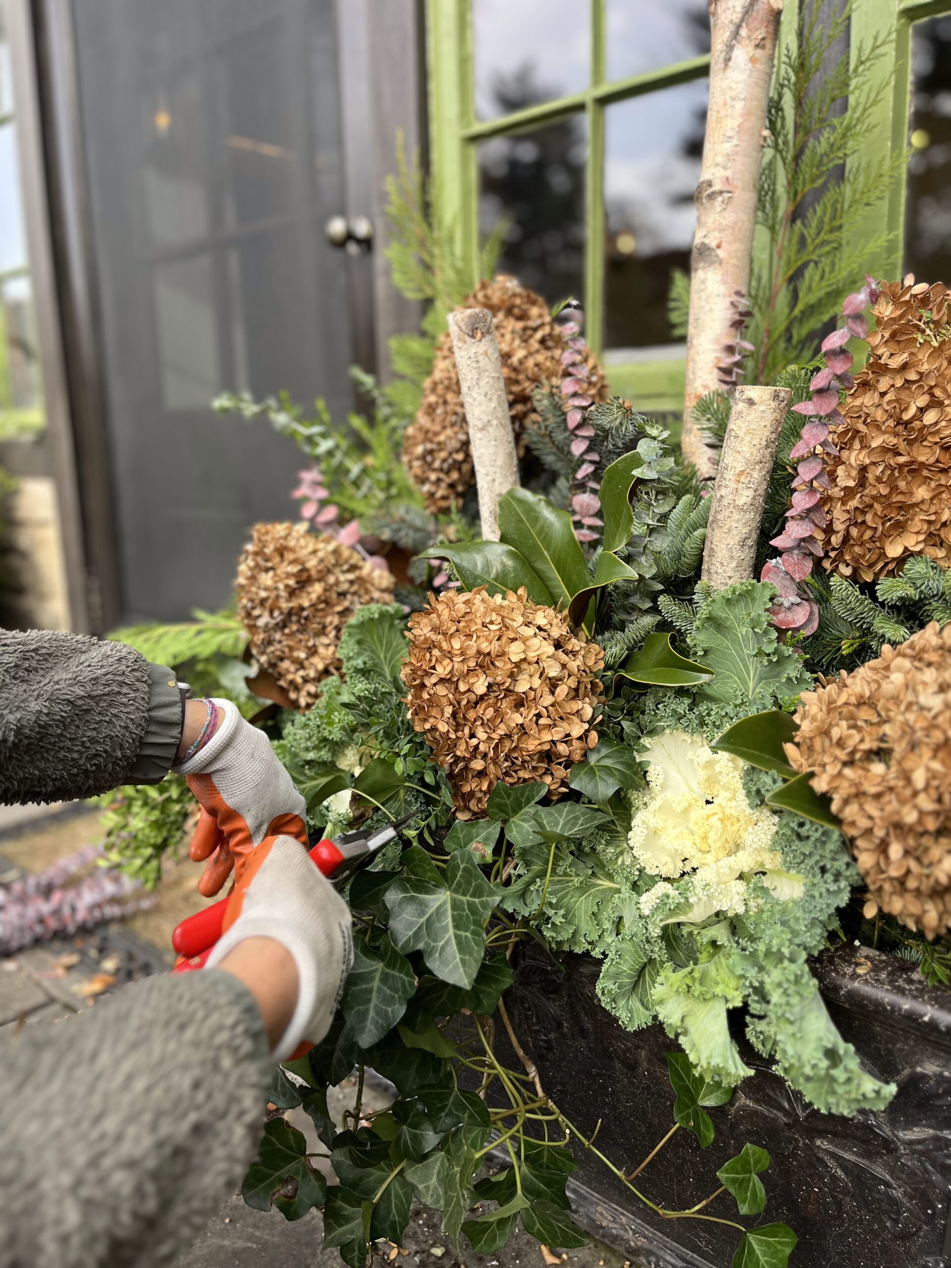 Person trimming outdoor flower arrangement by window.